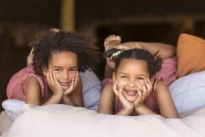Two young girls with curly hair resting on large cushions, smiling and posing for the camera.